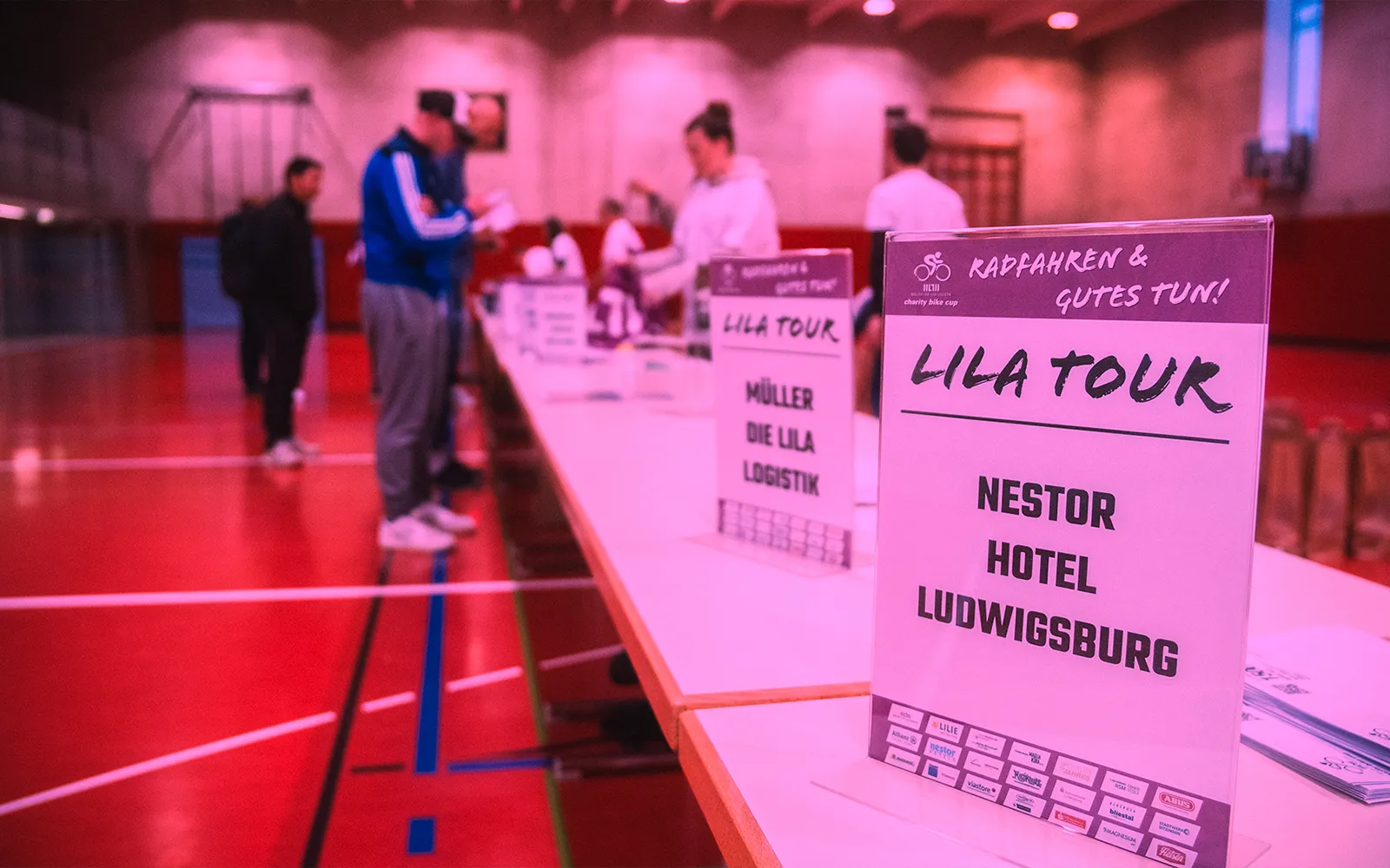 Startnummernausgabe in der Sporthalle mit LILA TOUR Schildern