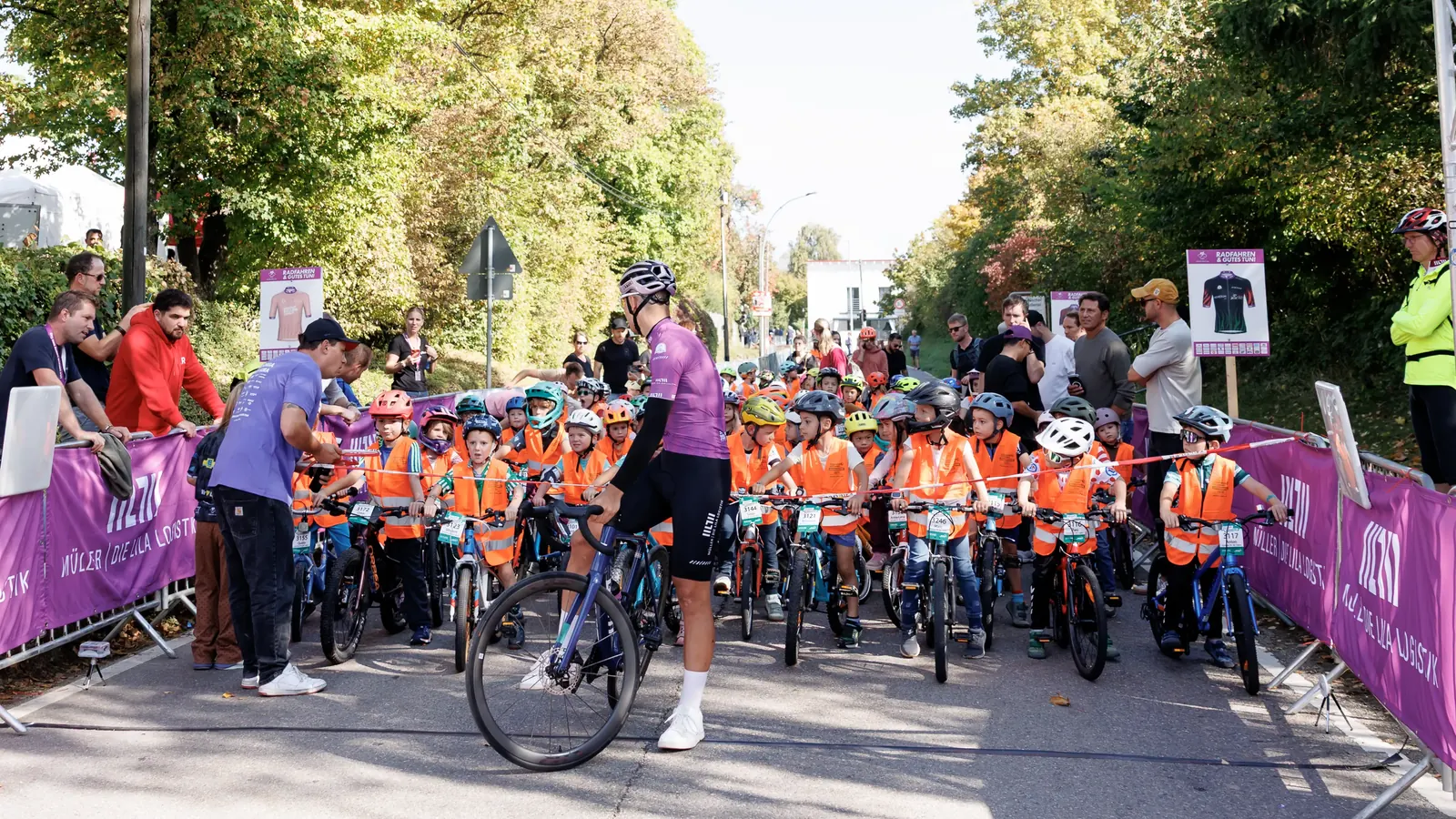 Startaufstellung der Kinderrunde: Kinder in orangen Warnwesten mit Fahrrädern hinter dem Startband