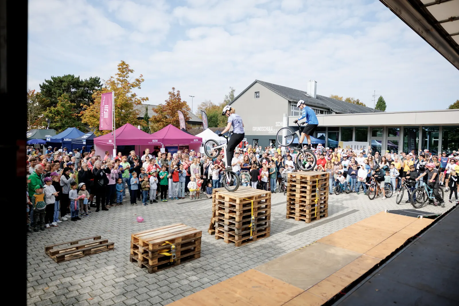 Trial-Show vor der Grundschule: zwei Fahrer balancieren auf Palettenstapeln vor dichtem Publikum